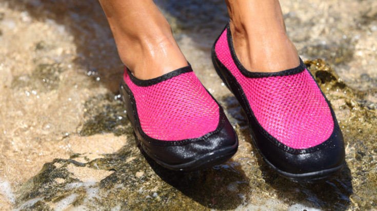 Water shoes / swim shoe in Pink neoprene on rocks in water on beach. Closeup detail of the feet of a woman wearing bright pink neoprene water shoes standing on rocks at the edge of the ocean.