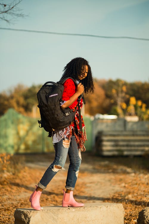 black model wearing striped blouse, red scarf, ripped jeans and pink rubber ankle boots carrying backpack